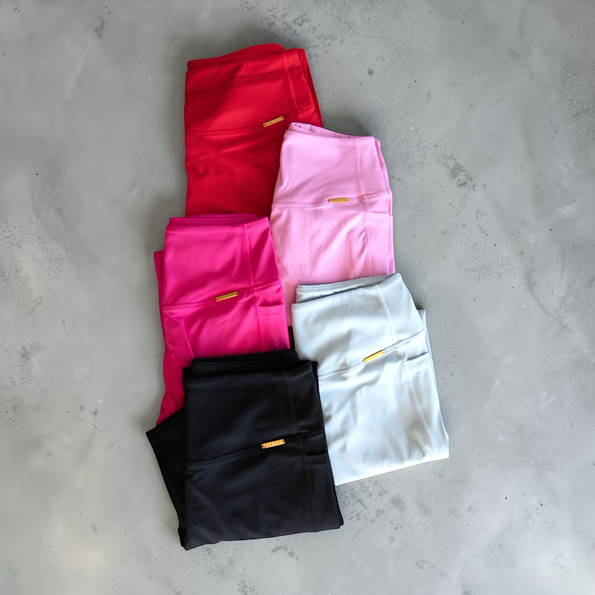 Four pairs of folded leggings in red, pink, black, and gray on a concrete floor.