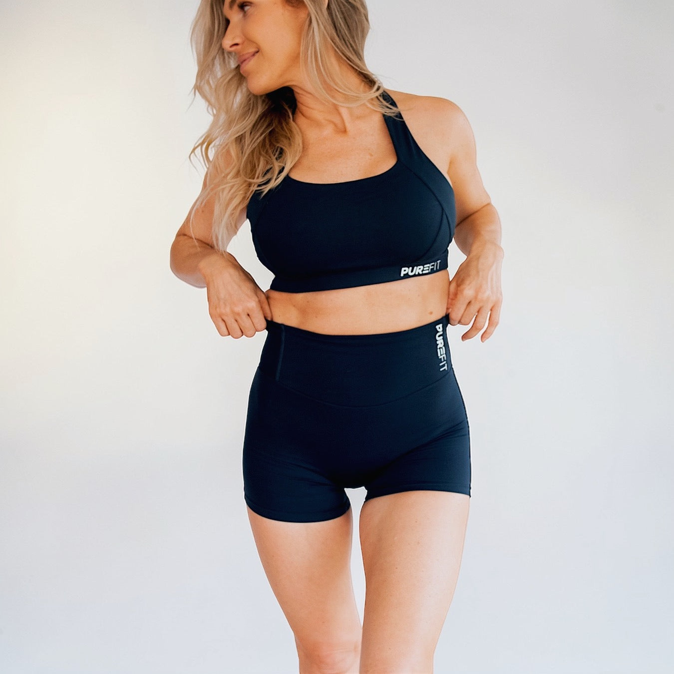 Woman wearing black sports bra and shorts with 'PUREFIT' branding on a light background