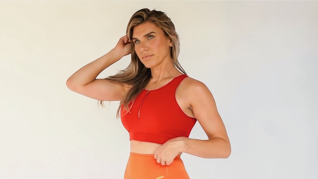 Woman wearing a red sports bra with a gold zipper feature on the front, standing against a neutral background